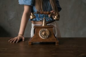 A woman in a blue blouse stands with a vintage wooden rotary phone on a wooden table.