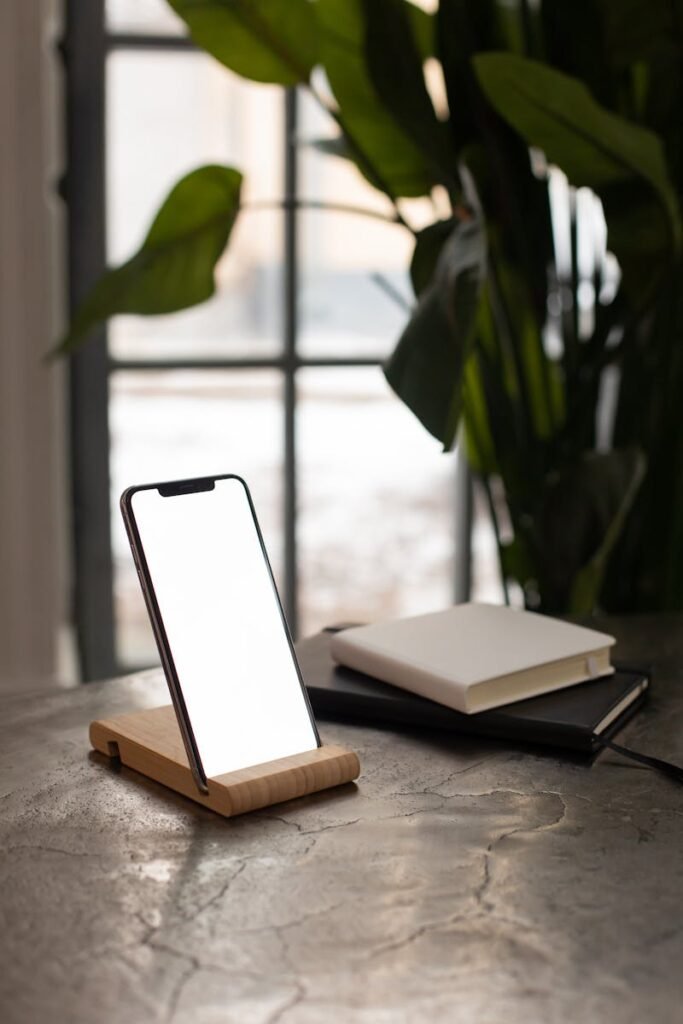 A smartphone on a wooden stand beside notebooks and a large plant indoors.