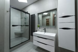 White sink at wall with mirror near cupboard in modern bathroom with glass shower cabin and bright illumination in apartment
