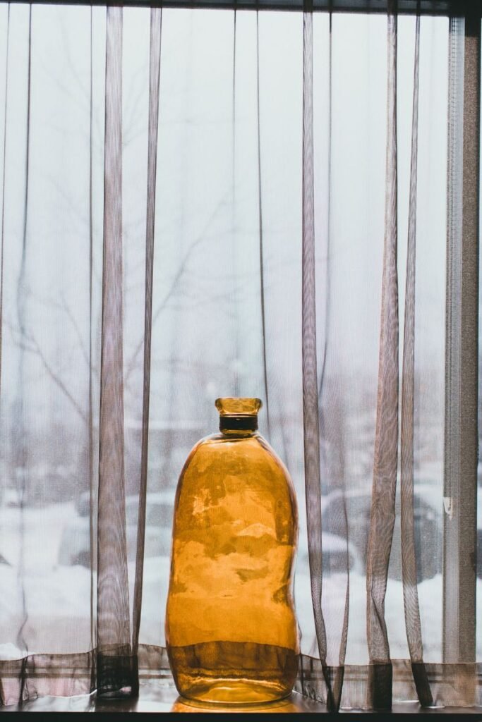 A vertical shot of an amber glass bottle against sheer curtains with a snowy outdoor backdrop.