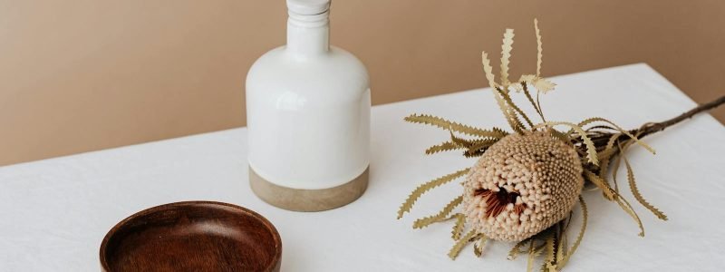 Elegant composition featuring a white bottle, wooden bowl, and dried flower on a table.