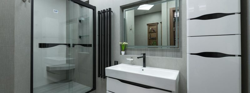 White sink at wall with mirror near cupboard in modern bathroom with glass shower cabin and bright illumination in apartment