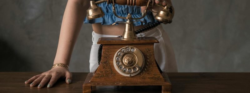 A woman in a blue blouse stands with a vintage wooden rotary phone on a wooden table.